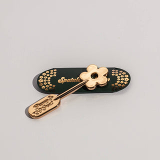 Gold metal lip care spatula with daisy-shaped applicator on decorated dark green case