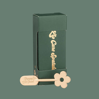 Green packaging box with gold daisy-shaped lip care spatula tool for cosmetic application.