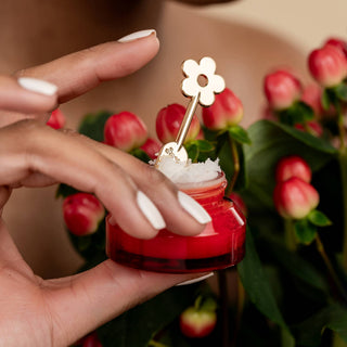 Hand holding red lip balm jar with gold daisy-shaped spatula tool surrounded by pink roses