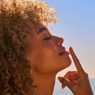 Woman with curly hair applying lip product with finger against sunset sky background