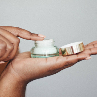Hands holding open glass jar of lip scrub with gold lid against neutral background