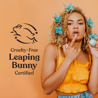 Woman applying lip balm with Leaping Bunny cruelty-free certification logo and orange background.