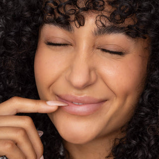 Woman with curly hair smiling with closed eyes, finger touching glossy tinted lips showcasing lip tint product.