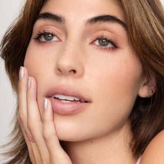 Woman with natural makeup wearing nude-pink lip tint, hand gently touching face showing manicured nails.