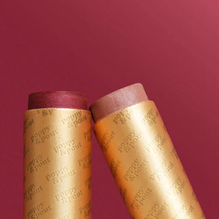 Two golden lip tint tubes with burgundy and pink caps against a red background.