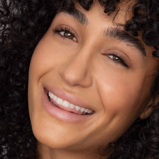 Smiling woman with curly dark hair wearing natural lip tint, showing healthy glowing skin and white teeth.