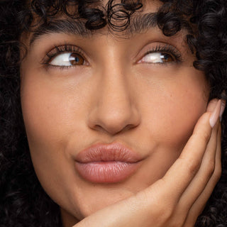 Woman with curly hair wearing natural pink lip tint, hand gently touching face.