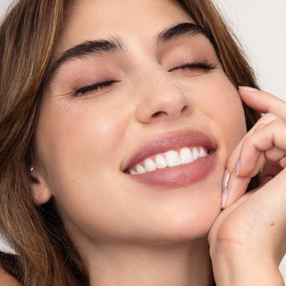 Woman with closed eyes smiling, showing white teeth, wearing natural pink lip tint with hand touching face.