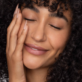 Woman with curly hair touching face wearing natural pink lip tint with closed eyes smiling