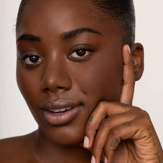 Woman with natural makeup showcasing nude lip tint with hand posed near face against neutral background.