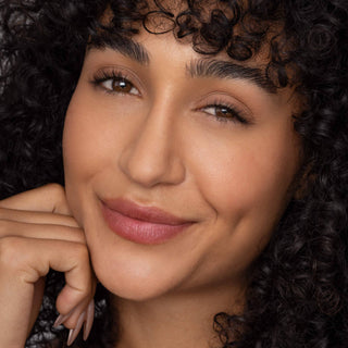 Woman with curly dark hair wearing natural lip tint, hand posed near face showing subtle makeup look.