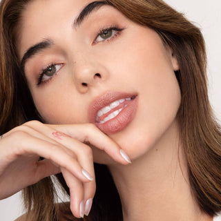 Woman with natural makeup wearing coral-pink lip tint, hand near face showing manicured nails.