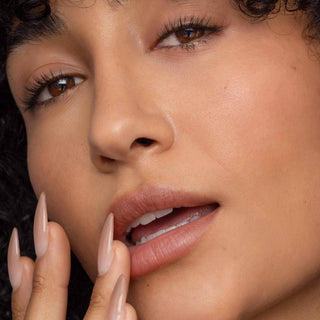 Woman with natural makeup touching her face, showcasing nude-pink lip tint and manicured nails.