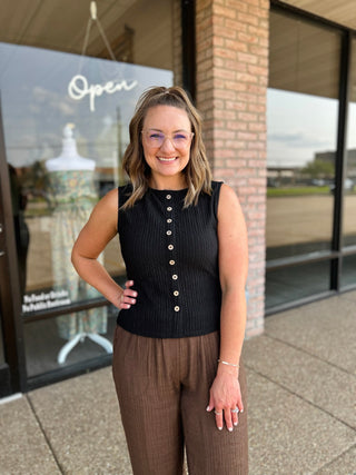 Woman wearing a sleeveless textured button-front top paired with wide-leg trousers outside a boutique.