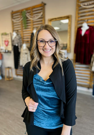 Woman wearing teal velvet tank top with black cardigan, smiling in boutique setting with clothing displays.