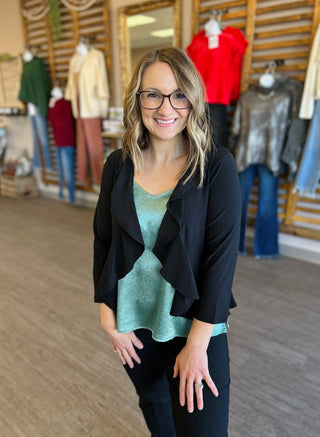 Woman wearing black ruffled cardigan over teal top in clothing store with mannequins displaying colorful apparel.