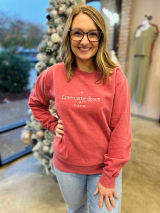 Woman wearing coral embroidered crewneck sweatshirt with "Love came down" text in retail store setting.