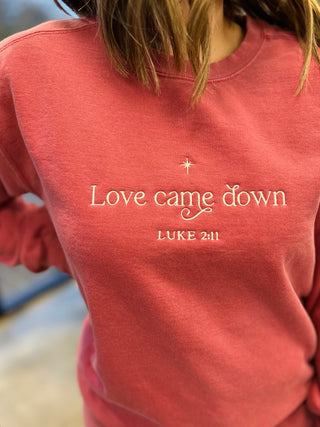 Woman wearing coral crewneck sweatshirt with "Love came down" and "LUKE 2:11" embroidered text and star design.