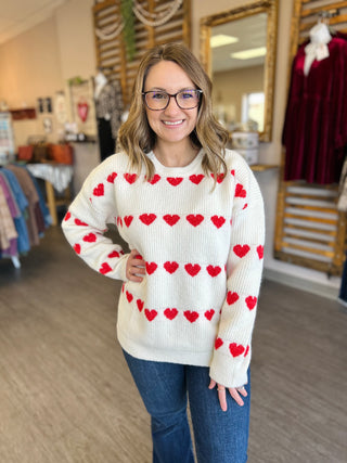 Woman wearing white knit sweater with red heart pattern, standing in clothing boutique store