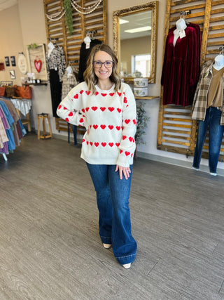 Woman wearing white heart-patterned sweater with jeans in clothing boutique store setting.