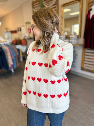 Woman wearing cream knit sweater with red heart pattern in retail store setting