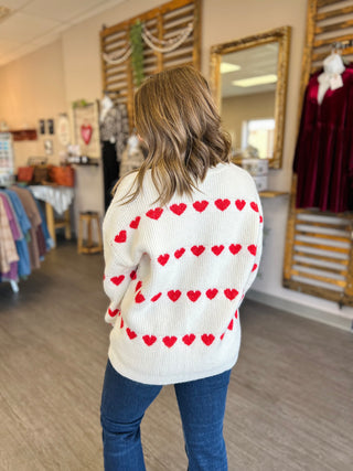 Woman wearing white knit sweater with red heart pattern, shown from behind in boutique setting.