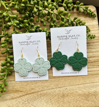 Harper Klay Co handcrafted clover-shaped clay earrings displayed on wooden surface with green succulent plants.