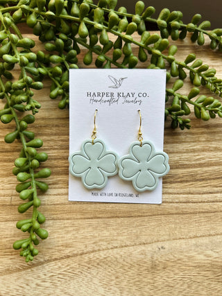 Light green clover-shaped earrings on product card surrounded by succulent plants on wooden surface.