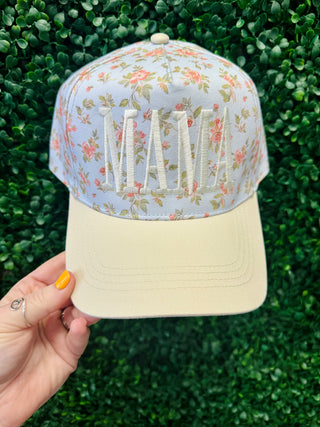 Hand holding MAMA embroidered floral pattern trucker hat with cream brim against green foliage background.