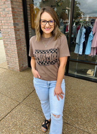 Woman wearing brown graphic mama t-shirt with distressed jeans standing outside clothing boutique storefront.