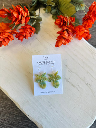 Handcrafted polymer clay leaf cluster earrings on gold hoops displayed on white wood with orange flowers.