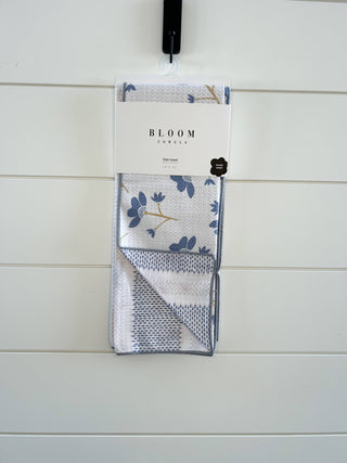 Bloom Towels dish towel in packaging hanging on white shiplap wall, featuring floral pattern and waffle weave texture.