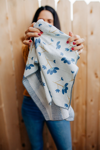 Woman holding floral patterned kitchen towel with blue botanical design against wooden fence background.