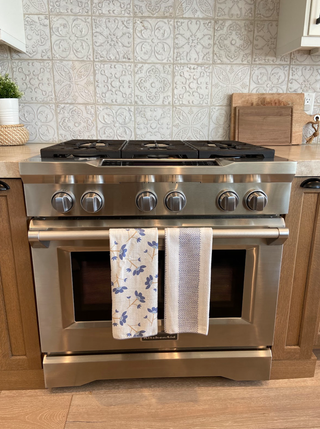 Kitchen towels hanging on stainless steel range with decorative tile backsplash and wooden cabinets