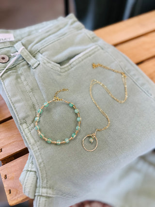 Gold chain necklace and green beaded bracelet displayed on light green denim jeans on wooden surface.