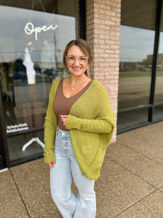 Woman smiling outside boutique wearing an open-front knit cardigan over a brown tank top with patterned jeans.