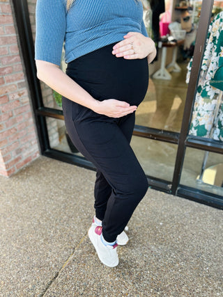 Pregnant woman wearing black maternity knit jogger pants with hands on belly, standing outdoors