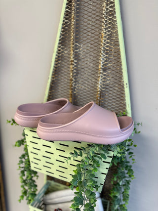Mauve platform slide sandals displayed on green basket with trailing plants in bright setting