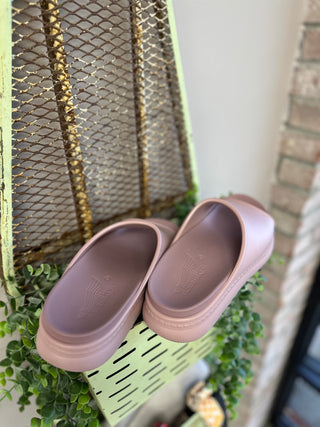 Mauve platform slide sandals displayed on green metal shelf with wire basket and plants