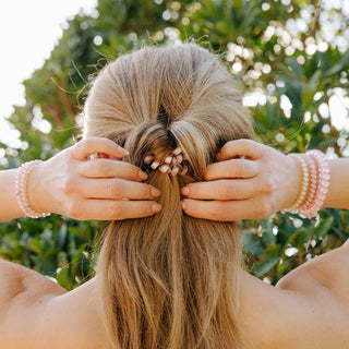 Woman styling blonde hair with decorative hair tie while wearing pearl bracelets outdoors