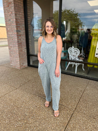 Woman wearing gray ribbed sleeveless jumpsuit with pockets, standing outside storefront in sandals