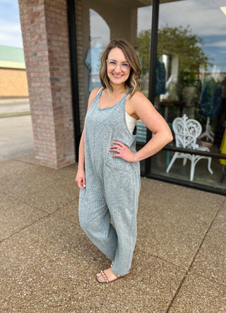 Woman wearing gray ribbed sleeveless jumpsuit standing outside storefront, smiling at camera.