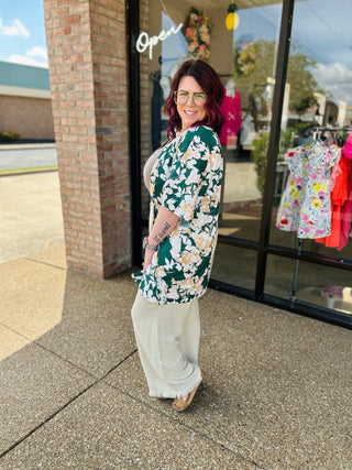 Woman wearing floral kimono over white pants standing outside boutique storefront, smiling at camera.