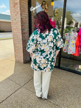 Woman wearing green floral kimono with white pants standing outside boutique storefront.