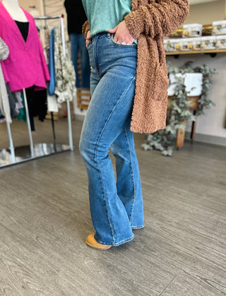 Person wearing medium wash high-rise flare jeans with brown cardigan in clothing boutique setting.