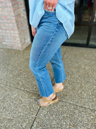 Person wearing medium wash straight leg jeans with raw hem and light blue sweater, walking on concrete sidewalk.