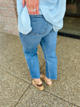 Woman wearing medium wash straight leg jeans with raw hem and light blue sweater, standing on concrete.