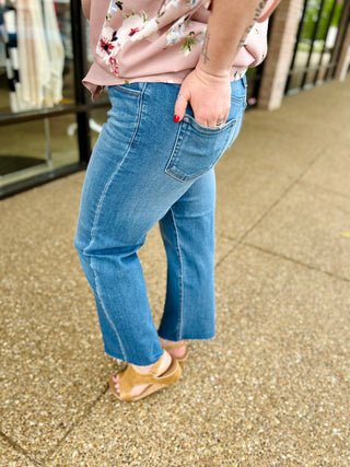 Woman wearing medium wash straight leg jeans with raw hem and floral top, hand in pocket.