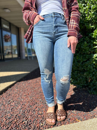 Person wearing distressed skinny jeans with raw hem and ripped knees, styled with plaid shirt and brown sandals.