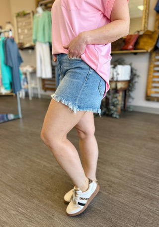 Woman wearing medium wash denim shorts with frayed hem and pink top in clothing store setting.